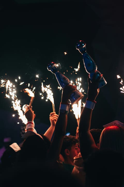 Raised hands hold two bottles high among lit sparklers in a dark, crowded club
